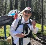 Maddie Aldrich with a backback, wearing a white shirt and baseball cap, hiking in the woods.